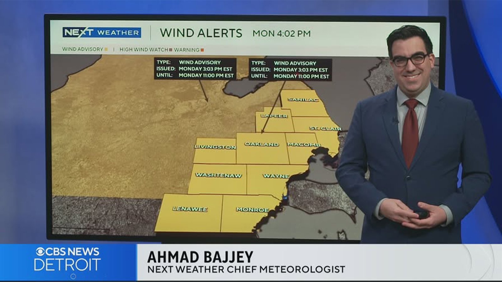 A man with dark hair wearing a suit and tie stands in front of a weather map on a TV screen that says wind alerts.