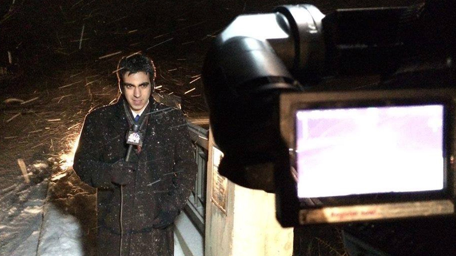 A man standing in the snow holds a microphone in front of a video camera.