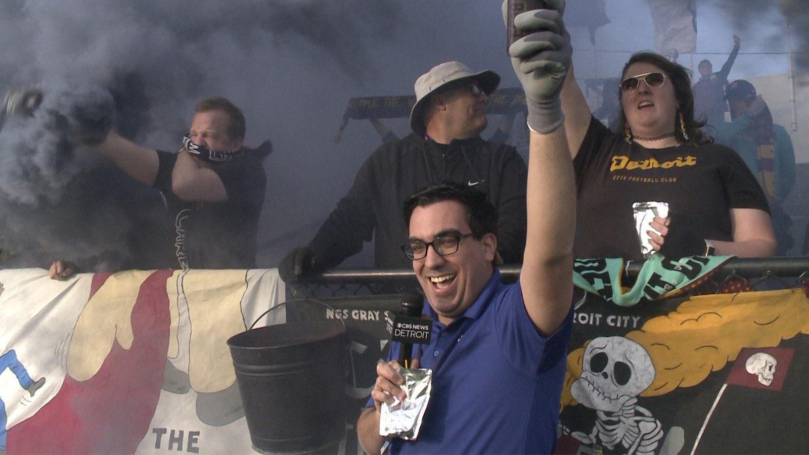 A man holds a microphone and smiles in front of fans who set off smoke bombs during a Detroit FC game.