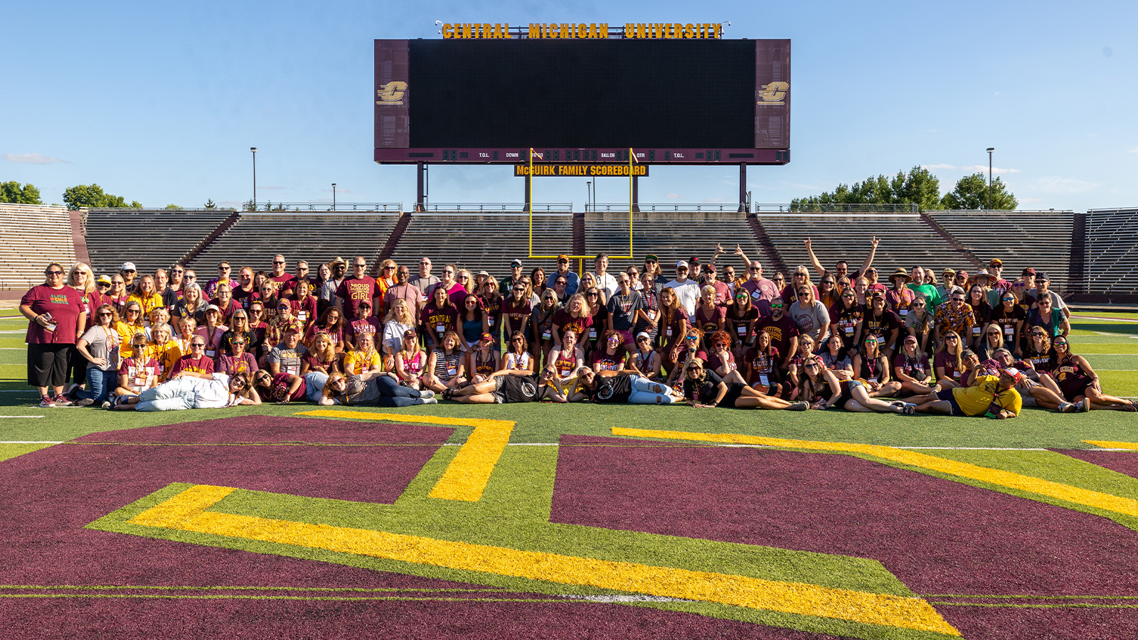 A large group of people pose for a photo on a football field in front of a jumbo scoreboard.