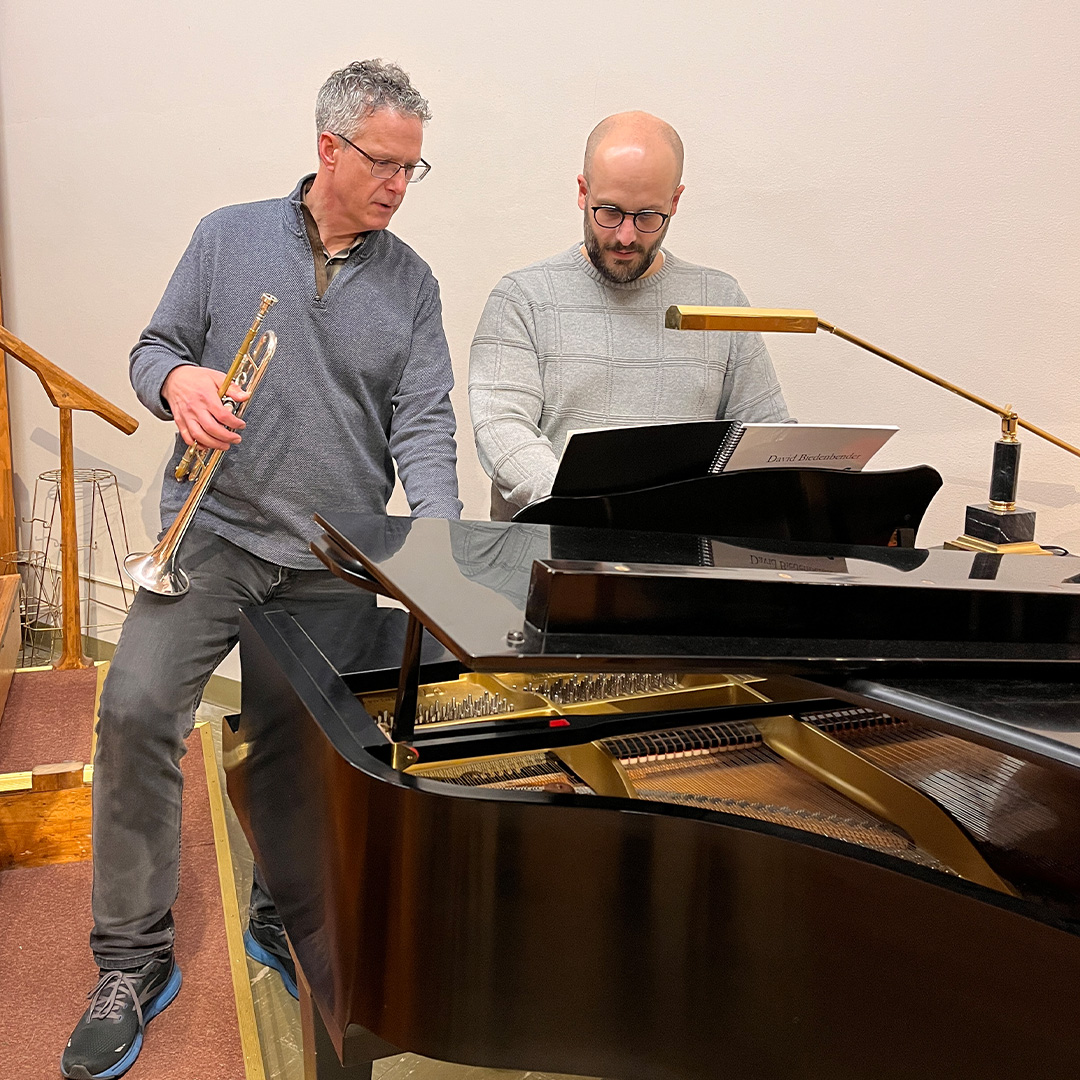 A man is holding a trumpet while standing next to a man who stands behind a piano.