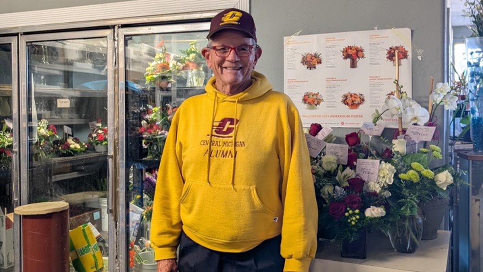 A man in a yellow Central Michigan Alumni sweatshirt with a maroon baseball hat is surrounded by flowers inside a flower shop.