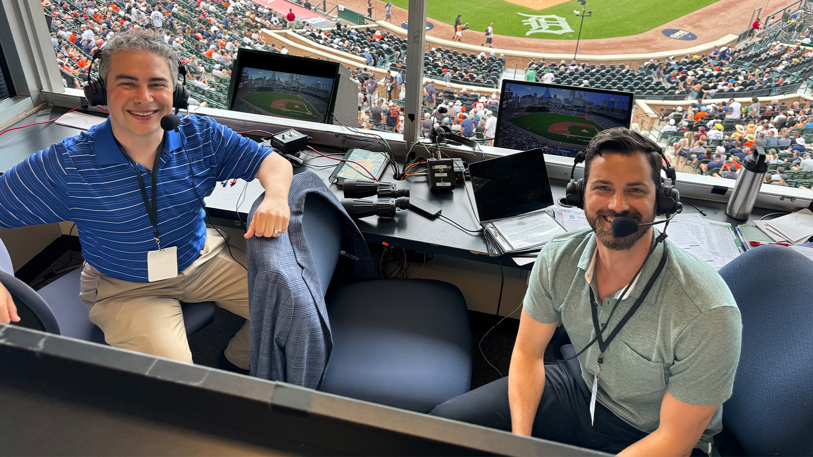 Two men sit in chairs in a stadium press box with headsets on and computers behind them.