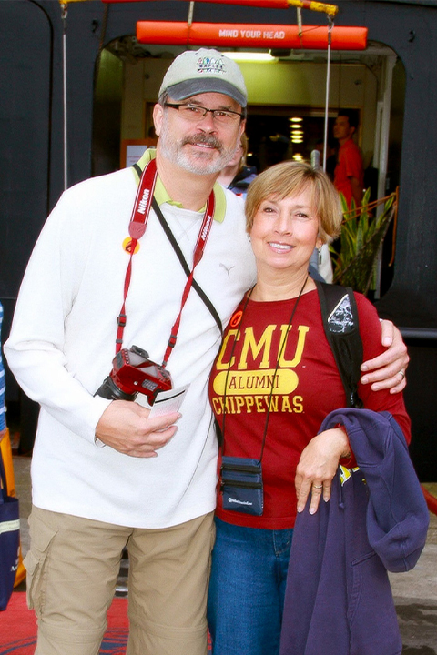 A man in a long sleeve white shirt and cargo khaki pants poses alongside a woman wearing a CMU Chippewas Alumni shirt and blue jeans.